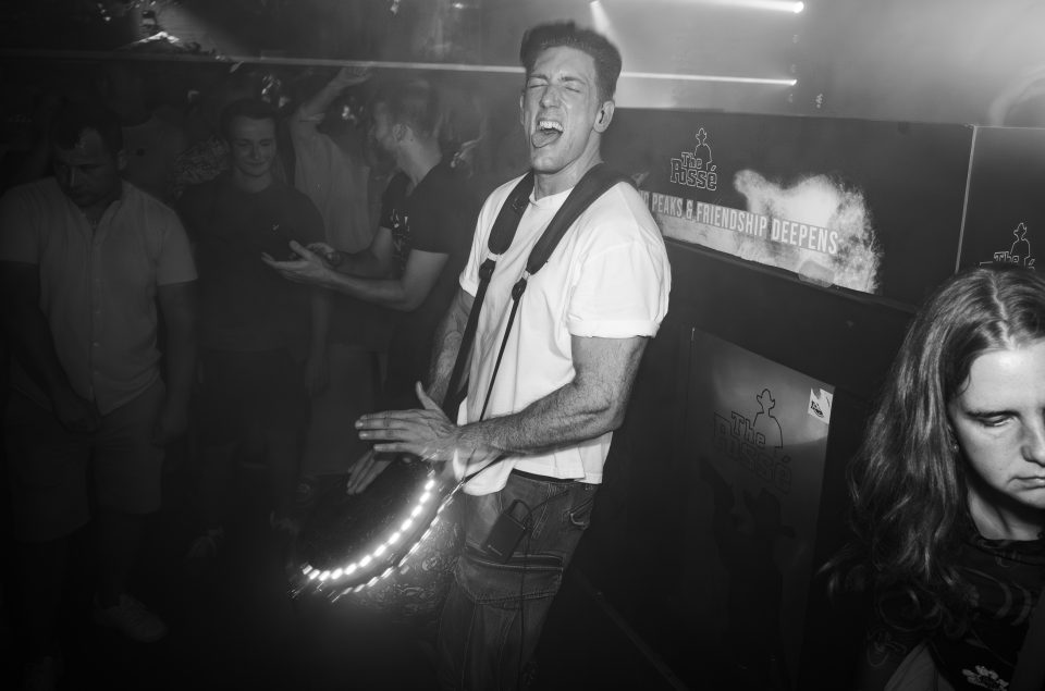 Black-and-white photo of a percussionist playing djembe with full energy in a club