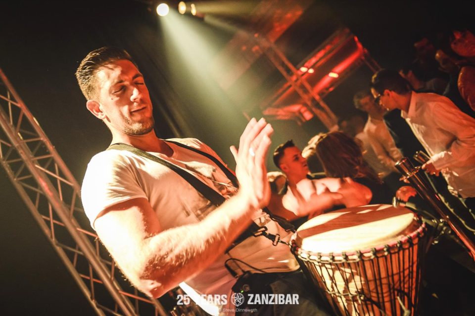 Percussionist playing djembe with eyes closed under a club spotlight