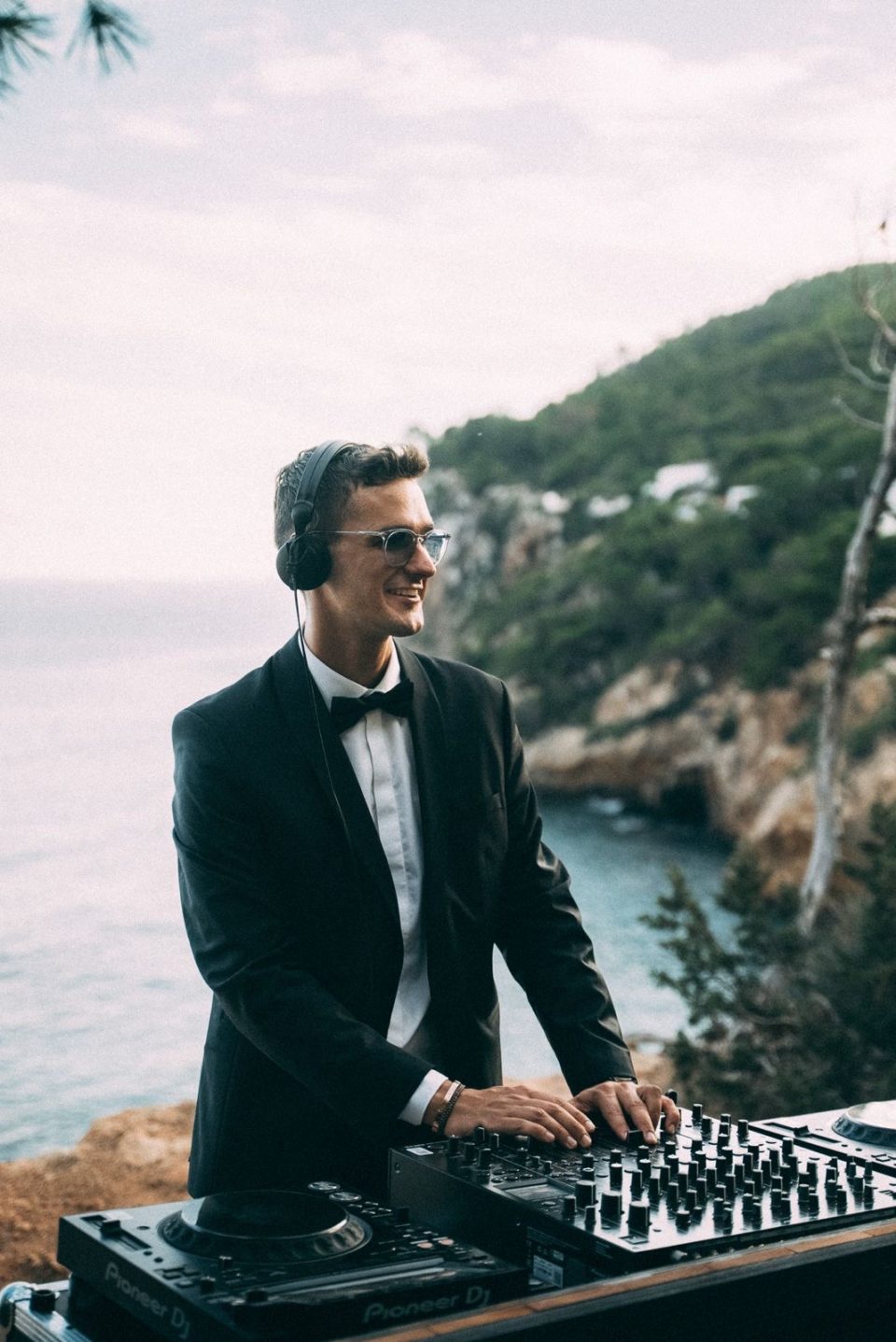 AMAXXY in a tuxedo playing a DJ set on a cliff above the sea