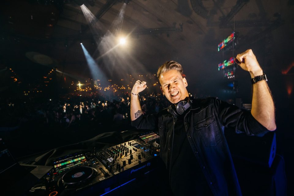 DJ Tom cheering with a raised fist behind the decks on a big stage